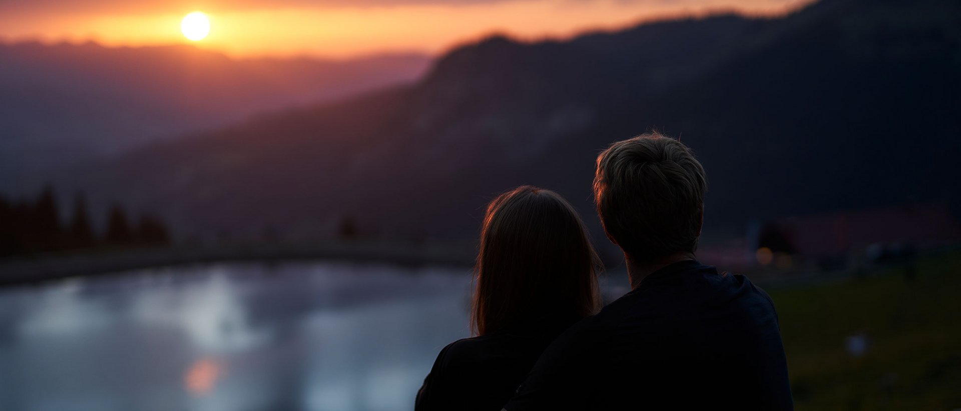 Panoramahotel Oberjoch | Bergbahnen Oberjoch Paar sitzt am See und betrachtet den Sonnenuntergang in den Bergen