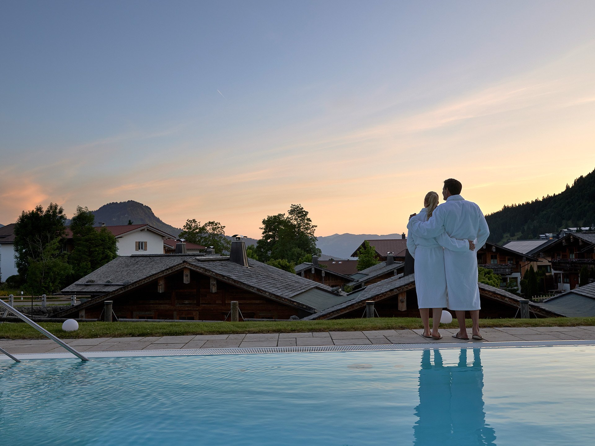 Entspannte Tage im 4*S Panoramahotel Oberjoch! Paar in Bademänteln blickt auf Berglandschaft bei Sonnenuntergang am Pool