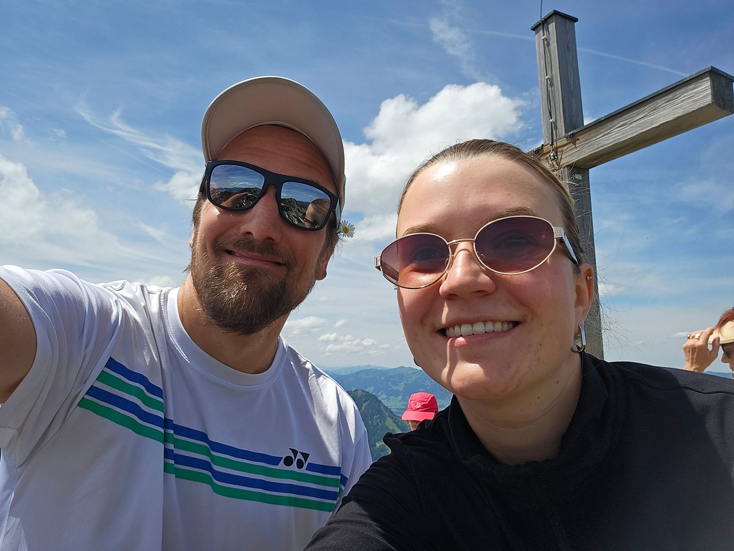 Exklusive Wandertipps von Basti Zwei lächelnde Personen mit Sonnenbrillen vor einem Holzkreuz auf einem Berg