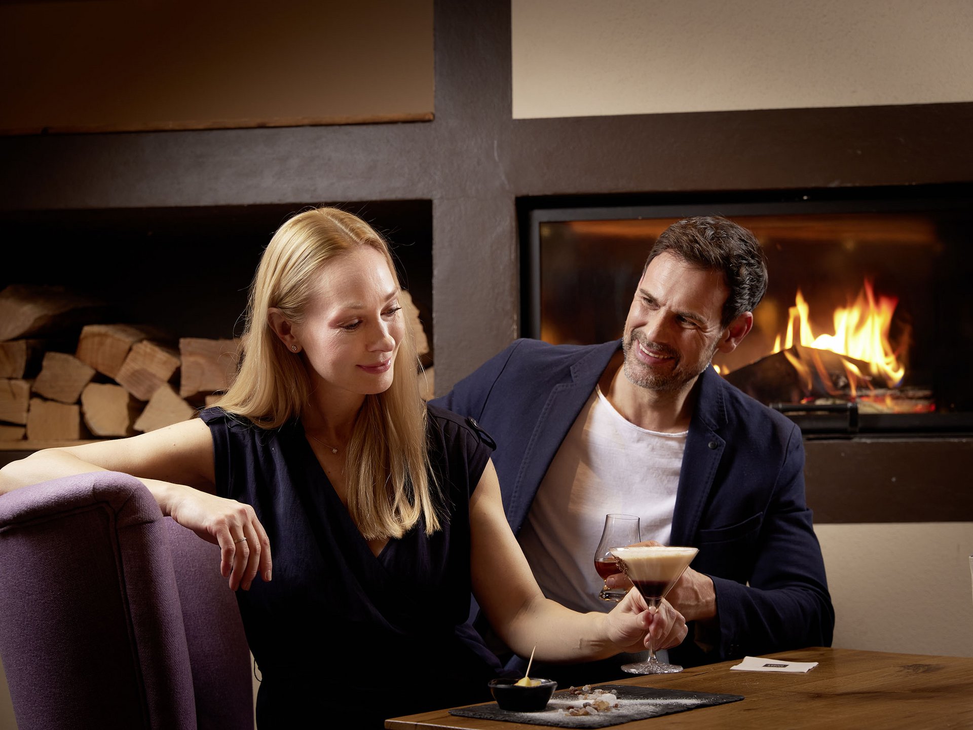 Entspannte Tage im 4*S Panoramahotel Oberjoch! Paar genießt Drinks vor Kamin in gemütlicher Lounge