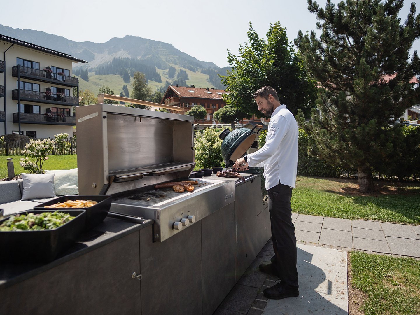 Panoramahotel Oberjoch | Küchenparty im Panoramarestaurant Mann grillt im Garten mit Bergen und Häusern im Hintergrund
